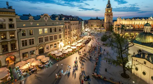 Wieczorny gwar na Rynku Głównym w Krakowie. Ludzie spacerują, siedzą w ogródkach kawiarnianych. W tle Sukiennice i wieża ratuszowa. To serce miasta, jego **sarbinowo centrum**.