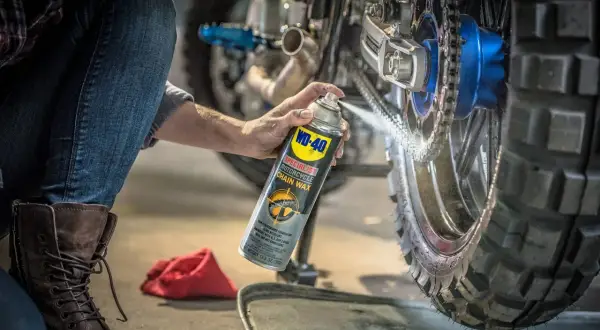 Persona aplica cera en aerosol WD-40 para limpiar la cadena de la moto, preparándola para la próxima aventura.