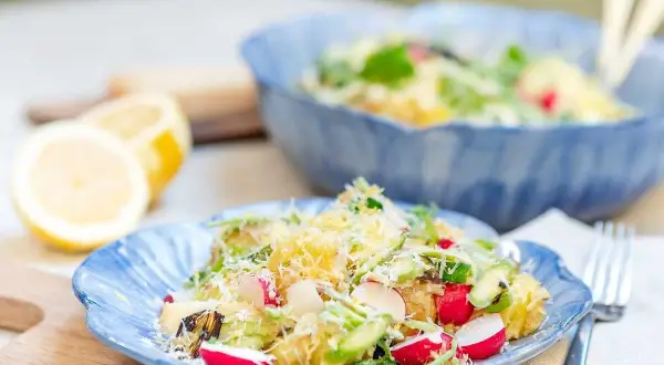 En härlig potatissallad med sparris, rädisor och hyvlad ost. Serveras på en blå tallrik med citroner i bakgrunden.