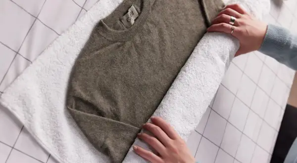 Hände rollen einen braunen Kaschmirpullover auf einem weißen Handtuch auf, um ihn nach dem Kaschmir waschen zu trocknen.