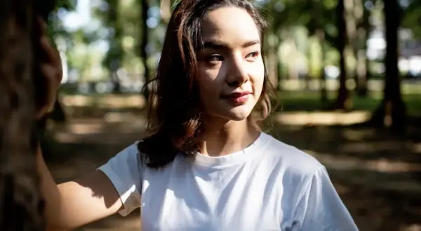 A young woman in a white t-shirt stands in a park, her face illuminated by dappled sunlight, showcasing the dramatic contrast of soft lighting vs hard lighting.