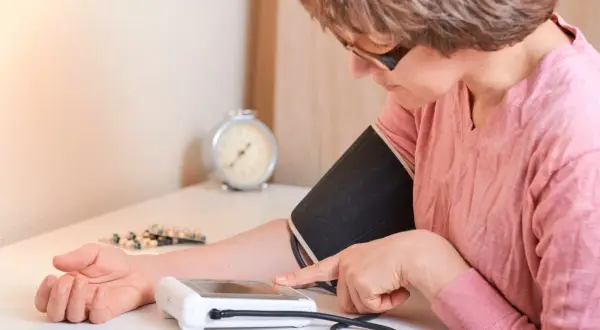 Kobieta mierzy ciśnienie, zmagając się z **wysokim ciśnieniem**. Na stole leżą leki.