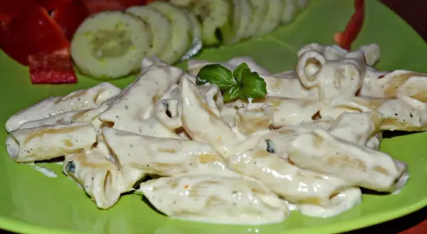 Penne w kremowym sosie z mozzarellą, udekorowane bazylią. Obok plasterki ogórka i papryka.