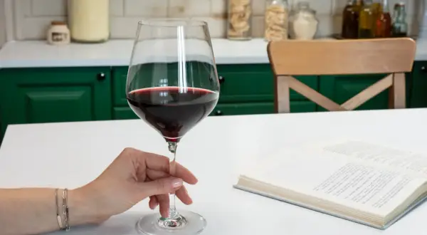 Dłoń trzyma kieliszek czerwonego wina, obok leży otwarta książka. Standardowy kieliszek ma ok. 150 ml.