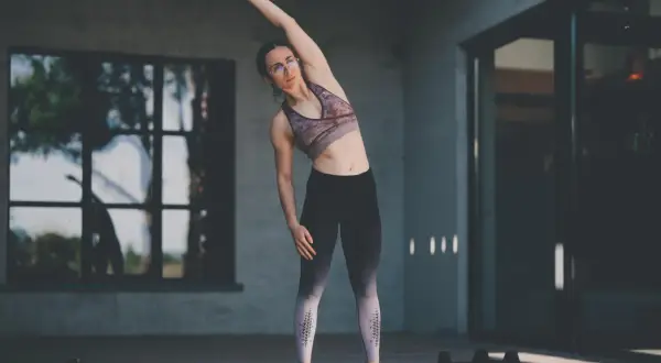 Femme faisant des étirements pour sa séance de musculation maison. Elle est en tenue de sport, bras levé, penchée sur le côté.