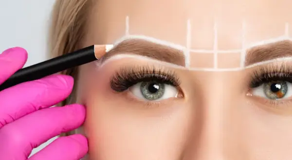 Manos enguantadas en rosa dibujan con lápiz blanco para crear cejas perfectas, marcando la simetría y el diseño.
