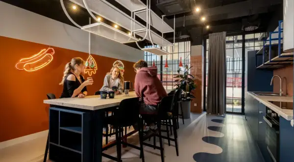 Studenci przy stole, rozmawiają i piją napoje. W tle neonowe hot dogi i pizza. To miejsce sprzyja nauce i relaksowi.