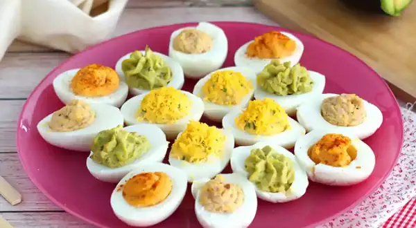 Assortiment d'œufs mimosa colorés sur une assiette rose. Des garnitures vertes, jaunes et orangées apportent une touche festive.
