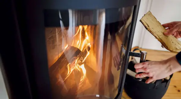 Hände legen Holzscheite in einen modernen Kamin, der brennt. Informieren Sie sich, welche Kamine noch erlaubt sind.