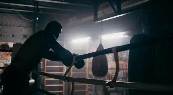 A boxer leans on the ropes in a dimly lit gym, contemplating how to get into boxing matches. Punching bags hang in the background.
