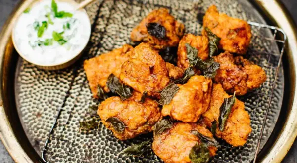 Knusprige chicken pakora, garniert mit frischen Kräutern, auf einem Gitterrost serviert. Daneben eine Schale mit Joghurt-Dip.