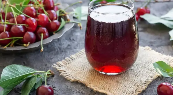 Szklanka orzeźwiającego soku z wiśni, obok miseczki pełnej świeżych owoców. Idealny na lato, prosty sok z wiśni przepis.