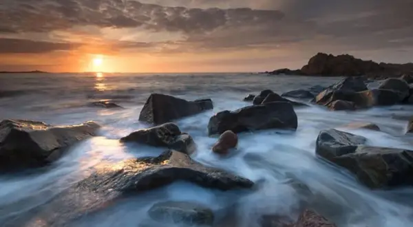Available light photos capture a serene sunset over a rocky coastline. Water flows gently around dark, wet stones, reflecting the warm hues of the sky.