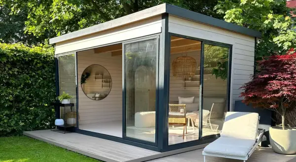 Modernes Gartenhaus mit Glasfront, das zum Streichen einlädt. Gemütliche Inneneinrichtung mit Rattankorb-Lampe und Liegestuhl draußen.