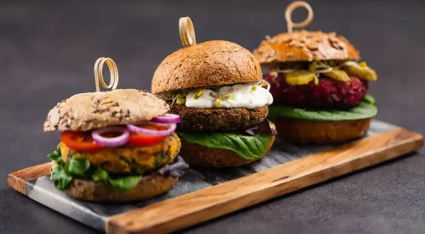 Drei appetitliche vegetarische Gerichte: Burger mit bunten Patties, frischem Gemüse und cremigen Saucen auf rustikalem Holzbrett.