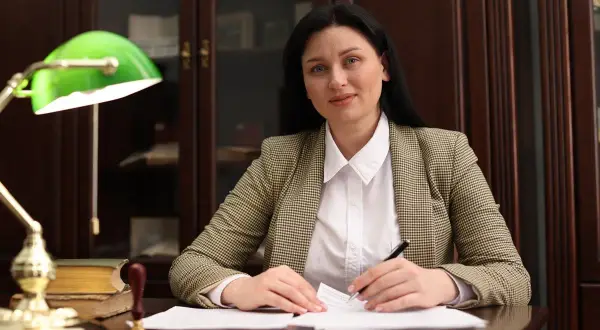 Kobieta w marynarce siedzi przy biurku, pisząc piórem na papierze, obok lampka i książki.