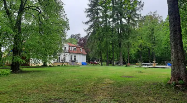 Biały dworek z czerwonym dachem otoczony zielenią. W oddali widać fontannę i drzewa. To urokliwa **gierłoż**.