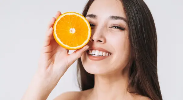 Lächelnde Frau hält Orangenscheibe vor ihr Auge. Vitamin C kann bei Rosacea helfen, die Haut zu beruhigen.