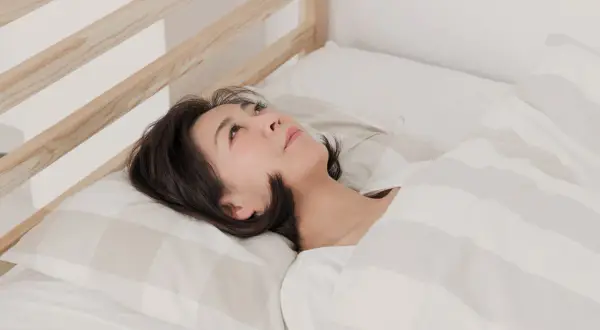 A woman lies in bed, looking up thoughtfully. Is it safe to sleep while dehydrated? Her expression suggests she's pondering this question.