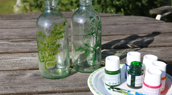 Två glasflaskor målas med gröna växtmotiv. Bredvid står färgtuber och penslar, redo att måla ljuslykta.
