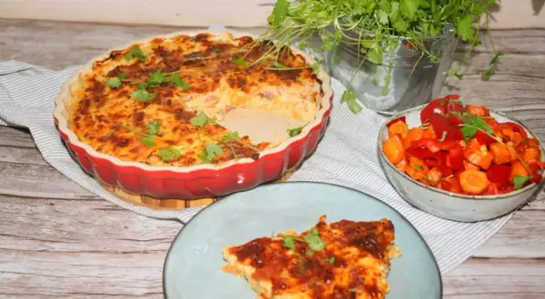 En gyllene paj med keso och en bit serverad på en tallrik. Bredvid står en skål med grönsaker och en kruka med örter.