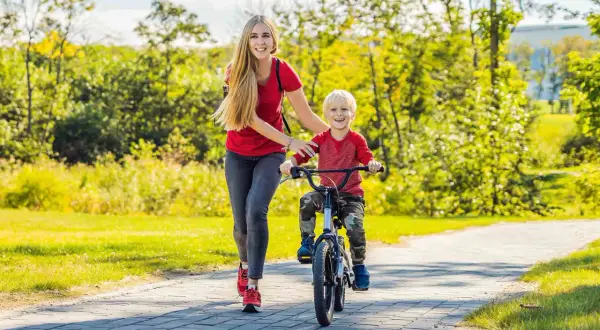 Mama uczy synka, jak nauczyć dziecko jeździć na rowerze. Radosny chłopiec pedałuje, a mama go asekuruje.