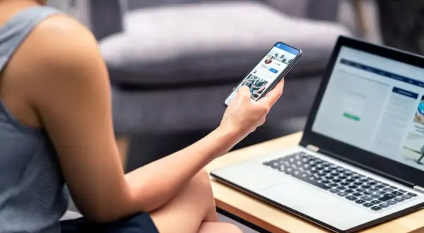 Kobieta przegląda Facebooka na telefonie, szukając informacji, jak wyłączyć status aktywności na FB. Obok leży laptop.