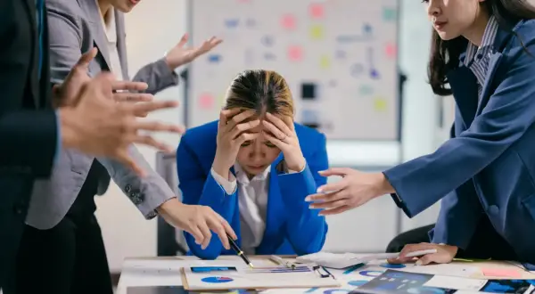 Giovane donna d'affari stressata, con le mani sulla testa, mentre colleghi la circondano con gesti agitati. Lo stress lavoro correlato è un rischio da valutare.