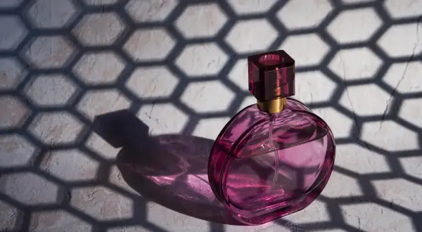 Różowa buteleczka perfum Dior J'adore, oświetlona słońcem, rzuca cień o geometrycznym wzorze plastra miodu.