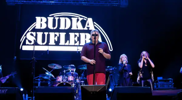 Budka Suflera na scenie. Wokalista w okularach śpiewa, a obok niego dwie wokalistki. W tle perkusja i logo zespołu.