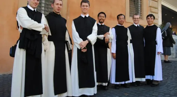 Siedmiu zakonników w białych habitach i czarnych szatach stoi przed pomarańczową ścianą.