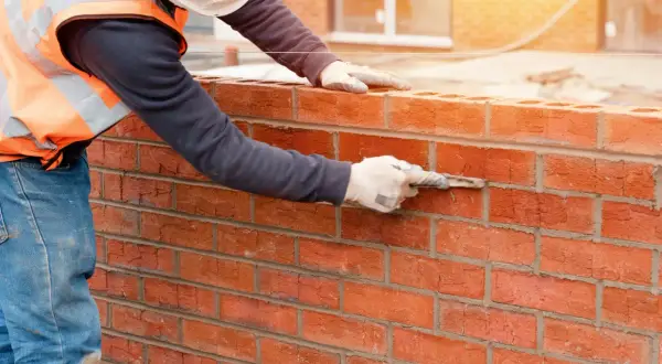 Muratore con guanti e cazzuola stende malta modellabile tra i mattoni rossi di un muro in costruzione.