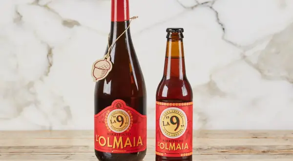 Due bottiglie di birra ambrata "La 9" del Birrificio L'Olmaia, una da 750ml e una più piccola, su un tavolo di legno.