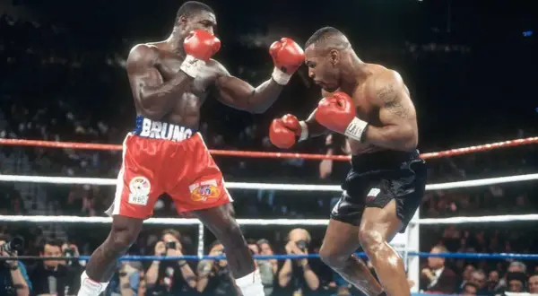 Two boxers, one in red shorts with "BRUNO" on the waistband, the other in black shorts, face off in a tense boxing match. The pressure fighter in red throws a punch.