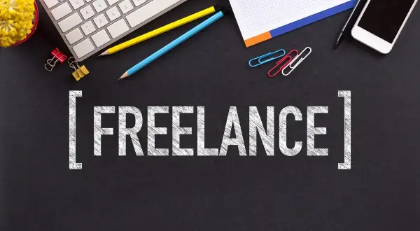 Biurko z klawiaturą, telefonem, notatnikiem i kredkami. Słowo "FREELANCE" na czarnym tle. To jest właśnie freelancer co to.