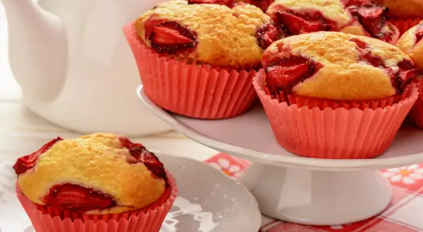 Pyszne muffiny z truskawkami w różowych papilotkach, gotowe do podania z białą czajniczkiem w tle.