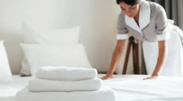 Pokojówka w hotelu dba o czystość i porządek. Na pierwszym planie stos białych ręczników, w tle kobieta poprawia pościel.