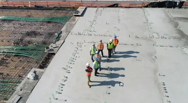 Grupa budowlańców na placu budowy, dyskutująca o tym, ile kosztuje kubik betonu. Widać zbrojenie i świeżo wylany beton.