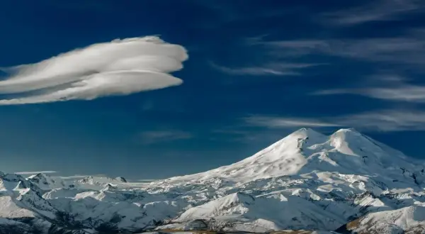 Pokryty śniegiem najwyższy szczyt Kaukazu, Elbrus, góruje nad ośnieżonymi pasmami górskimi pod błękitnym niebem z charakterystyczną chmurą.