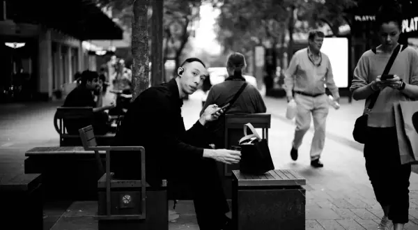 A man with earbuds looks at his phone while sitting on a bench. This 35mm vs 50mm street photography captures a moment of urban life.