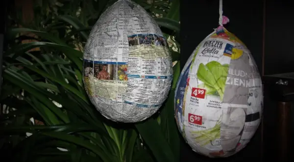 Jak zrobic jajko z gipsu w balonie? Dwa papierowe jajka, jedno z gazet, drugie z reklam, wiszą na tle zielonych liści.