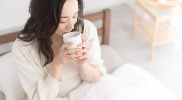 Donna con capelli scuri beve acqua calda al mattino, seduta a letto, avvolta in una coperta bianca.