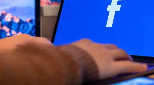 Dłoń na klawiaturze laptopa, na ekranie logo Facebooka. Dowiedz się, jak utworzyć stronę na Facebooku.