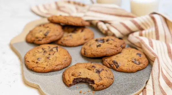 Nybakade chocolate chip cookies recept på en grå platta, med en bit tagen ur en cookie. Två glas mjölk och en randig handduk i bakgrunden.