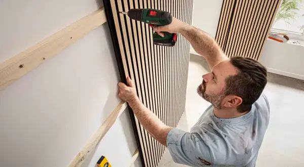Mann bringt mit Akkuschrauber vertikale Lamellen an der Wand an. Das Anbringen von Deckenpaneelen ist ein DIY-Projekt.