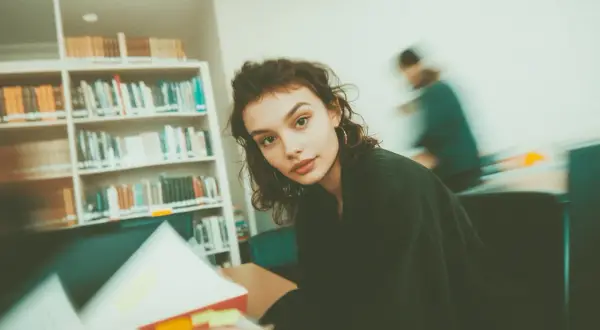Młoda kobieta w okularach, otoczona książkami, studiuje w bibliotece. Ibiden, nauka i wiedza.