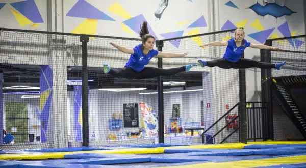 Dwie kobiety w niebieskich koszulkach wykonują szpagat w powietrzu na trampolinach.