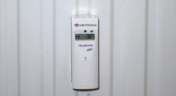 Biały, cyfrowy ciepłomierz Metrona Telmetric star przymocowany do żeberka grzejnika. Wyświetla temperaturę 38.7.