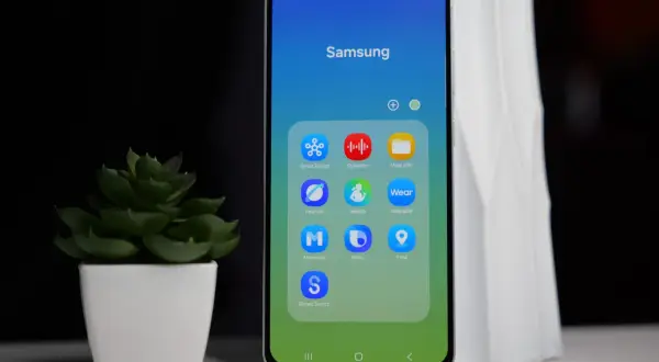 Telefon Samsung z ikonami aplikacji, w tym Bixby. Obok stoi zielona roślina w białej doniczce.