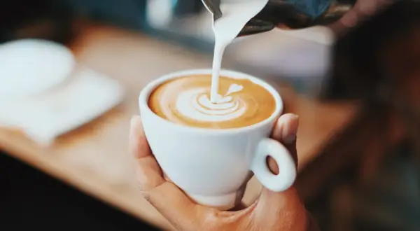 En hand håller en vit kopp med en vacker kaffe latte, där mjölken skapar ett hjärta i skummet.